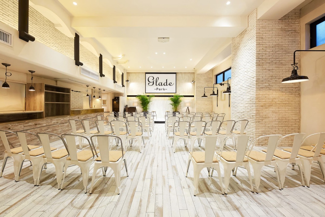 GLADE PARK 恵比寿の大広間。白を基調とした明るい宴会場に、アイアン製の白いチェアがシアター形式で並び、奥の壁には「Glade Park」のロゴサインが掲げられたパーティー会場の内観。